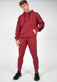 Bordeaux hoodie met zwarte strepen en logo, gecombineerd met bijpassende tapered joggers. Materiaal lijkt zacht met een gladde textuur en zakken.