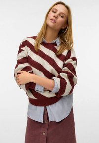 Gestrickter Pullover mit bordeauxroten und cremefarbenen diagonalen Streifen, lockere Passform und gerippte Bündchen, über einem hellblauen Hemd mit Knöpfen getragen.