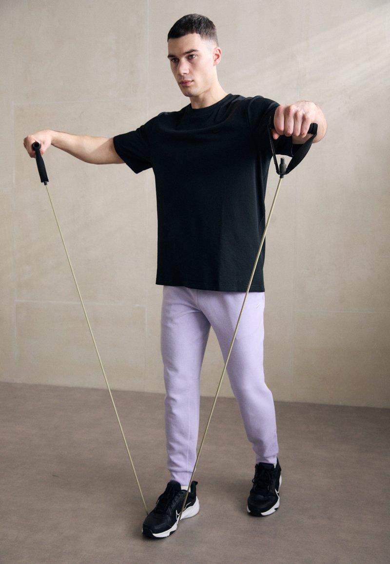 Widerstandsbänder in Gebrauch von einer Person, die ein schwarzes T-Shirt und helllila Jogginghosen trägt, mit schwarzen Sportschuhen. Glatte Textur, funktionales Design.