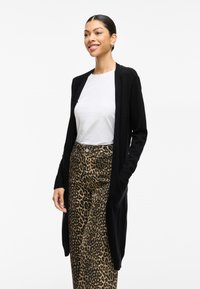 Svart lång kofta med öppen front, ribbade muddar och sidofickor, bärs över en vit skjorta och leopardmönstrade byxor på en modell.