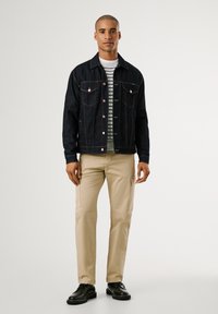 Chaqueta de mezclilla oscura con dos bolsillos en el pecho, camiseta de rayas blancas y grises debajo, pantalones cargo beige y zapatos negros.