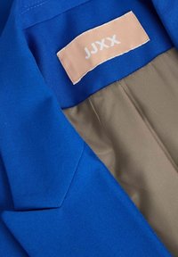Blå tygblazer med krage och en slät beige innersida. Har en krämfärgad etikett med "JJXX" tryckt i vitt.