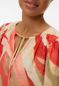 La blouse présente une base crème avec des motifs abstraits rouges et pêches vifs. Manches bouffantes, col rond et un détail en goutte d'eau avec une fermeture à nouer.