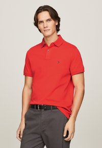 Röd polo t-shirt i bomull med klassisk krage, korta ärmar och en liten logotyp på bröstet. Bar med grå byxor och ett svart bälte.