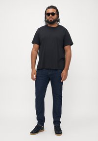 Mann med rastafletter, iført solbriller, sort t-skjorte, blå jeans og sorte joggesko, som står foran en ensfarget hvit bakgrunn.