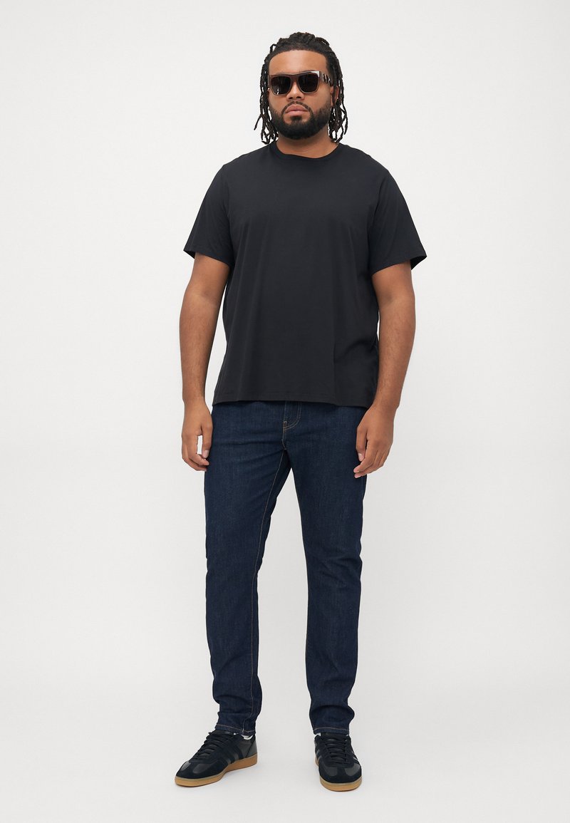 Mann mit Dreadlocks, der Sonnenbrillen, ein schwarzes T-Shirt, blaue Jeans und schwarze Turnschuhe trägt und vor einem schlichten weißen Hintergrund steht.