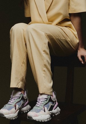 Persona sentada en una silla de madera oscura, vistiendo pantalones beige y zapatillas gruesas y coloridas con detalles en tonos pastel y cordones.