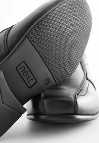 Zapato de cuero negro con suela de goma texturizada. Presenta un acabado suave, costuras elevadas y una marca en el talón.