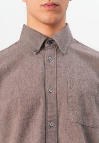 Hombre con una camisa marrón de botones, con cuello puntiagudo y un bolsillo en el pecho, visto desde el cuello hasta la mitad del torso.