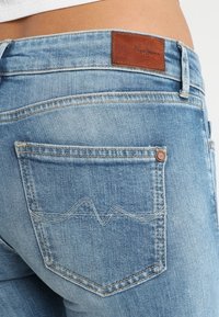 Los jeans de denim azul cuentan con una etiqueta de cuero en la cintura. Los bolsillos traseros presentan un sutil bordado en un patrón ondulado.