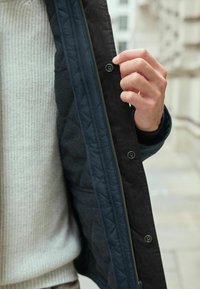 Gepolsterte, dunkelblaue Jacke mit einer strukturierten grauen Fütterung. Verfügt über metallene Druckknöpfe und einen Reißverschluss. Ein hellgrauer Pullover ist sichtbar darunter.