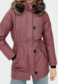 Veste matelassée rose avec capuche bordée de fourrure, boutons noirs, poches latérales et taille ajustable avec cordon. Tissu à texture lisse.