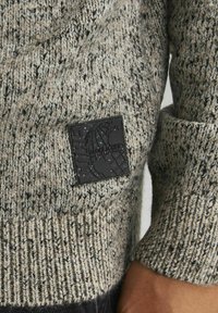 Sweter z dzianiny w odcieniach szaro-beżowych, z teksturowanym wzorem oraz czarną łatą z wytłoczonym logo na lewym przedzie.