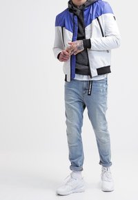 Homme portant une veste bleu et blanc par-dessus un hoodie gris, un jean clair déchiré retroussé au niveau des chevilles, des baskets blanches et des tatouages visibles sur les mains.