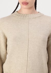 Beige stickad tröja med ribbad rund halsringning, med en central söm och strukturerad tyg. Mjuk, mysig look med långa ärmar.