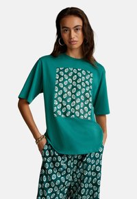 Tyrkysové oversize tričko vyrobené z měkké bavlny má velký vzorovaný čtverec s bílými designy listů. Slučuje se s odpovídajícími vzorovanými šortkami.