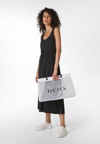 Borsa a rete bianca con la scritta "DEHA", abito senza maniche nero con vita regolabile e sneaker bianche, in piedi su uno sfondo neutro.
