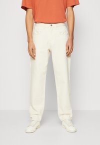 Men's cream-colored pants made of cotton, featuring a straight fit, five pockets, and a button closure. Paired with a rust-colored T-shirt and white sneakers.