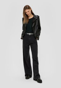 Schwarze Lederjacke mit Reißverschlüssen, dunkelgrünes Oberteil und hochgeschnittene schwarze weit geschnittene Jeans. Dicke schwarze Plateau-Stiefel runden das Outfit ab.