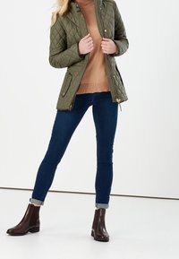 Gequiltede olijfgroene jas met een hoge kraag, gedragen over een bruine trui, gecombineerd met donkerblauwe jeans en bruine enkellaarsjes.