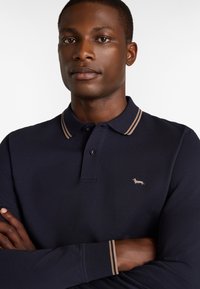 Navyblauer Langarm-Polohemd mit beigem gestreiftem Kragen, kleinem gesticktem Hundelogo auf der Brust und Knopfverschluss. Aus Baumwollmaterial.