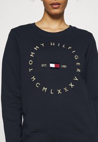 Sudadera azul marino con bordado dorado que presenta texto en forma circular y un pequeño diseño de bandera. Tejido suave, cuello redondo clásico y ajuste relajado.