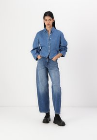 Giacca di denim di un blu chiaro con colletto, chiusura a bottoni e maniche oversize, abbinata a jeans blu a vita alta e scarpe con plateau nere.