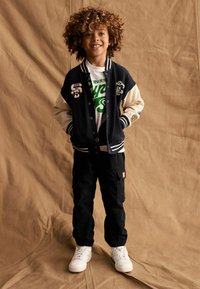Marineblaues und cremefarbenes Varsity-Jacket mit Aufnähern, weißes Grafik-T-Shirt, schwarze Cargo-Hosen und weiße Sneakers. Der Hintergrund ist aus strukturiertem beigem Stoff.
