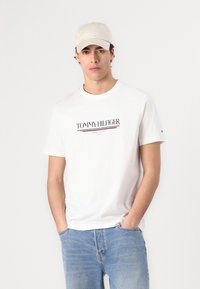 Bijela pamučna majica s okruglim izrezom, s mornarsko plavim i crvenim logotipom "TOMMY HILFIGER". Uparena sa svijetloplavim trapericama i bež kapom.