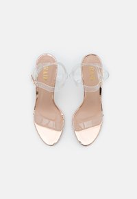 Paire de sandales en or rose à bout ouvert avec des brides transparentes à la cheville et aux orteils, vues de dessus sur un fond blanc.