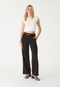 Weißes, kurzes Oberteil mit kurzen Ärmeln, schwarze hochgeschnittene Hosen und schwarze, gestreifte Absatzsandalen. Einfaches Design, figurbetonte Silhouette und glatte Texturen.