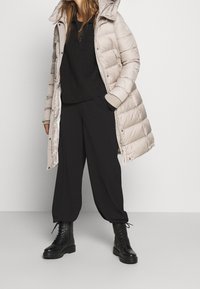 Lichtbeige pufferjack met capuchon, zwart gebreid trui en losse zwarte broek, gecombineerd met zwarte veterschoenen voor de strijd.