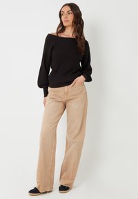 Zwarte geribbelde trui met een off-shoulder ontwerp, gecombineerd met lichtbeige wijde broekspijpen. Het model draagt zwarte schoenen met beige accenten.