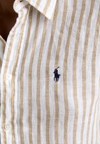 Beige és fehér vízszintes csíkos vászon ing, bal mellen sötétkék hímzett logóval és gombos gallérral.