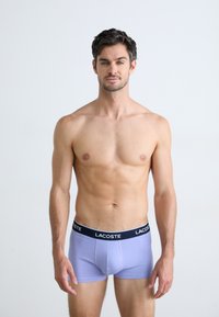 Lila boxershorts met een zwarte Lacoste tailleband, met een getailleerd ontwerp en zachte textuur, gedragen door een staande mannelijke model.