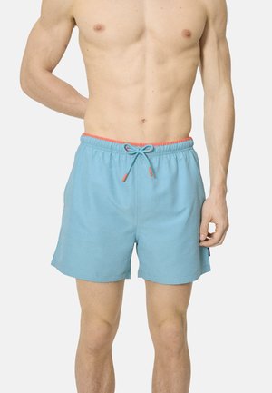 Hombre sin camisa vistiendo pantalones cortos de baño azul claro con cintura coral y cordón, de pie frente a un fondo liso.