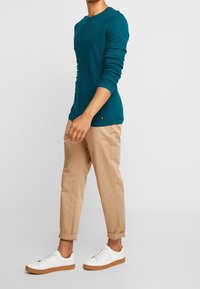 Turkos tröja med ribbat mönster, rund halsringning och långa ärmar, tillsammans med sandfärgade avsmalnande byxor och vita sneakers med gummisular.