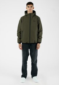 Giacca con cappuccio verde oliva con cerniere nere, polsini elastici e orlo con coulisse, abbinata a jeans neri e sneakers bianche.