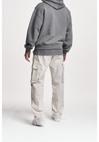 Šedá mikina s kapucí a volnějším střihem, spárovaná se krémovými cargo kalhotami s bočními kapsami a strukturovanou tkaninou. Bílá obuv dotváří vzhled.