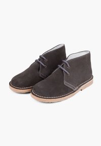 Botas desert de ante negro con textura suave, puntera redonda, dos ojales para los cordones y una suela de goma marrón. Diseño minimalista y artesanía visible.
