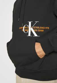 Černá mikina s kapucí, vybavená přední kapsou, stahovacími šňůrkami a výrazným bílým a oranžovým nápisem, který uvádí značku a rok jejího založení.