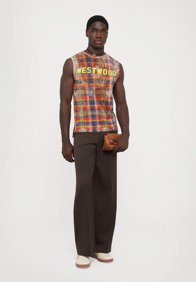 Man in een multicolore mouwloze Westwood-plaid top, donkere brede broek, witte schoenen, zwarte kralenketting, en houdt een kleine bruine tas vast.
