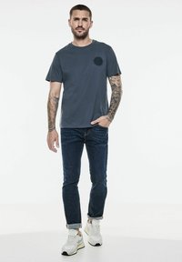 Mörkgrå t-shirt med kort ärm och rund halsringning, med en cirkulär svart detalj på bröstet, i kombination med mörkblå jeans och sneakers.