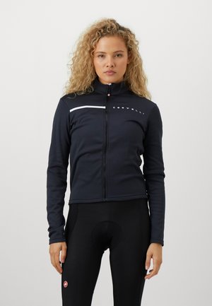 Frau mit lockigem blonden Haar, die eine schwarze Radjacke mit Reißverschluss und schwarze gepolsterte Radshorts trägt, steht vor einem einfarbigen Hintergrund.