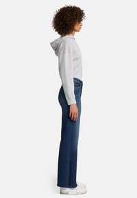 Felpa grigia con cappuccio e spalle scese, abbinata a jeans blu scuri a vita alta. Il modello indossa sneakers bianche. Vista di profilo.