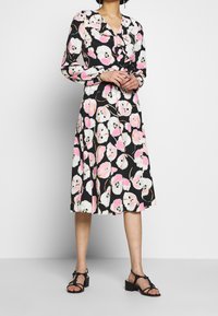 Svart klänning med rosa och vita blommönster, långärmad och med V-ringning. Kjolen är knälång och kombineras med svarta sandaler.