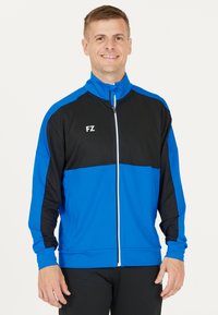 Blå och svart zip-up sportjacka med hög krage, utrustad med vit dragkedja och logotyp. Tillverkad av mjukt material med långa ärmar.