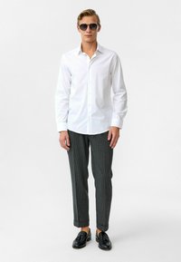 Witte button-up shirt, grijs gestreepte broek en zwarte lakschoenen. Man met zwarte zonnebril, staand met handen langs het lichaam op een effen achtergrond.