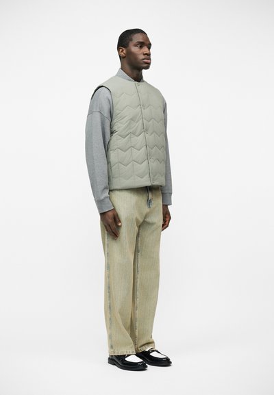 Homme debout portant une veste matelassée gris clair par-dessus une chemise à manches longues grise, un pantalon beige à jambes larges, et des chaussures noires et blanches sur fond blanc.