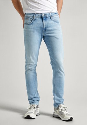 Homme portant un jean slim bleu clair et des baskets blanches, debout les mains dans le dos sur un fond neutre.